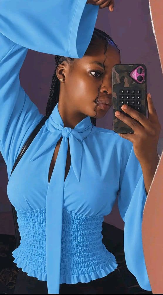 Haut bleu ciel à manches longues flottantes avec col noué et taille smockée porté par une femme prenant un selfie.
